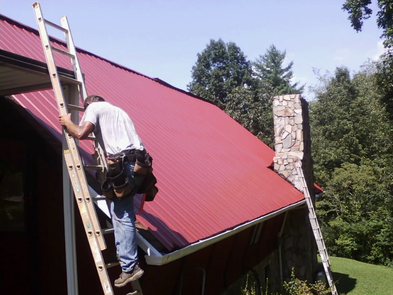 Roofer on a ladder beside a red metal roof and stone chimney — Soffit & Fascia Repair by Roofing Brothers in Thousand Palms
